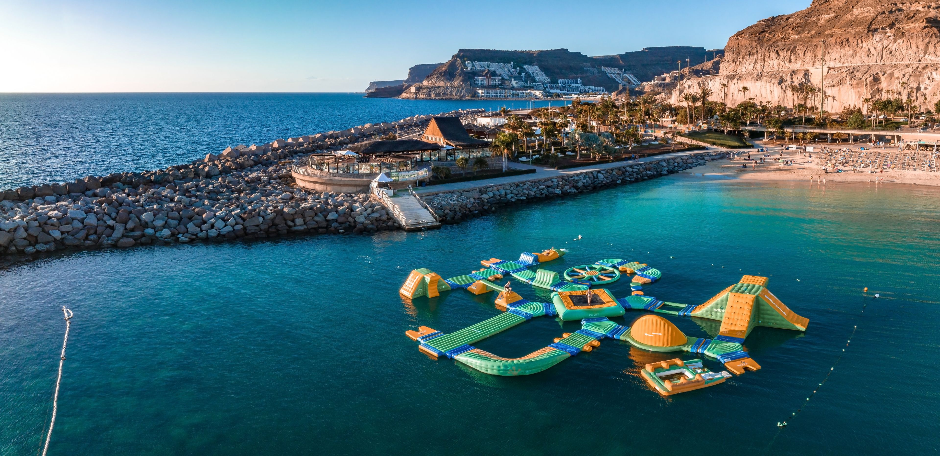 A coastal area with a rocky breakwater, a floating water park, and a mountainous backdrop.