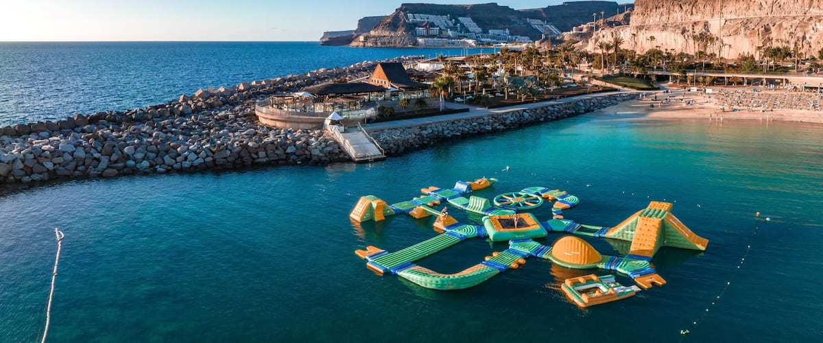 A coastal area with a rocky breakwater, a floating water park, and a mountainous backdrop.