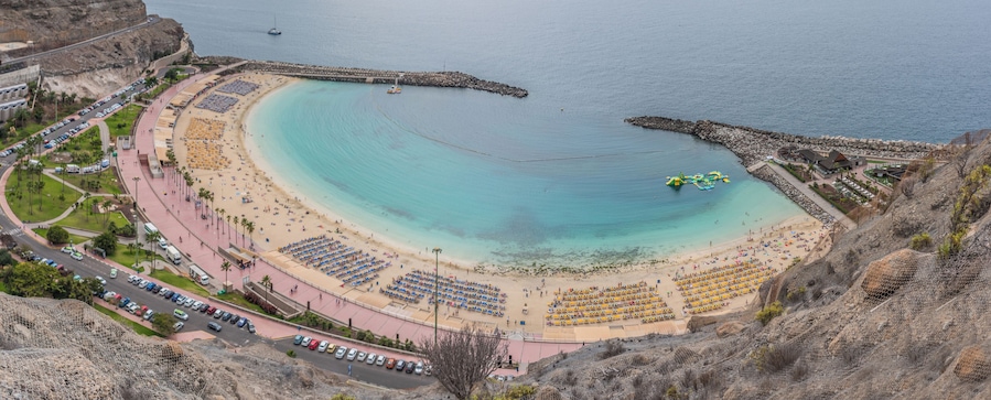 A coastal beach with a curved shoreline, a rocky breakwater, and a parking area with rows of cars.