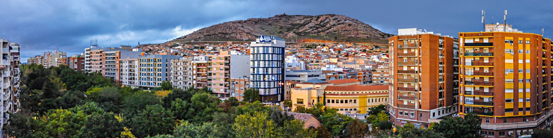A cityscape with a prominent hill in the background, surrounded by residential buildings and lush greenery.