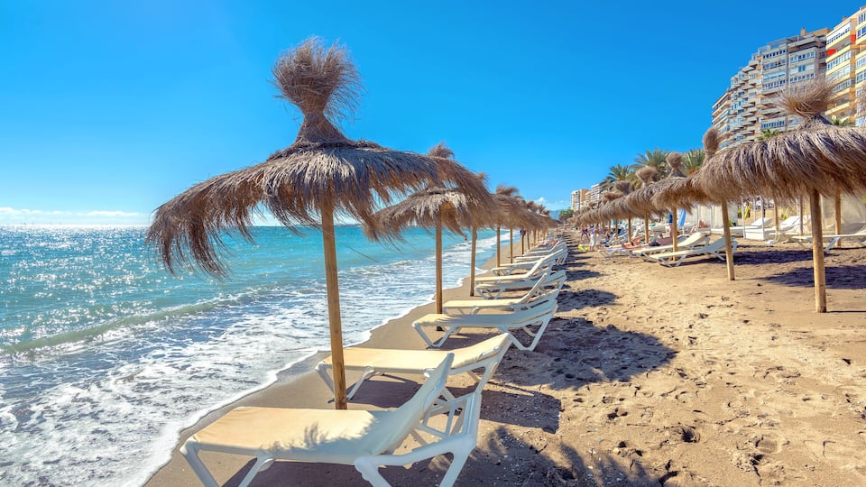 A beach with rows of sun loungers and thatched umbrellas, a clear blue sky, and ocean waves.