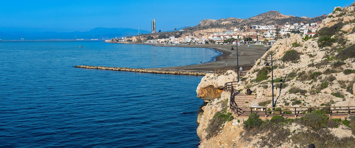 A coastal town with a rocky shoreline, a pier extending into the sea, and a prominent tower in the distance.