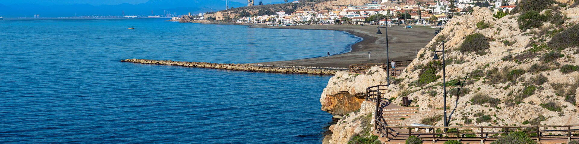 A coastal town with a rocky shoreline, a pier extending into the sea, and a prominent tower in the distance.