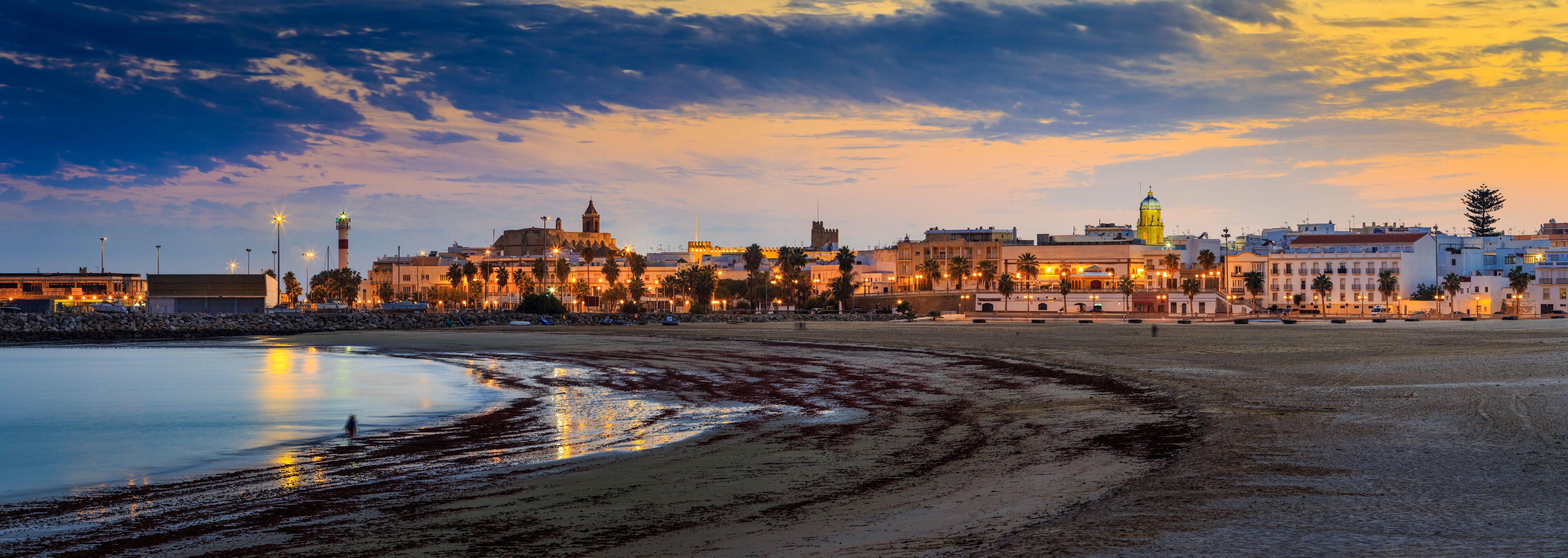 A coastal cityscape at dusk with illuminated buildings and a beach.