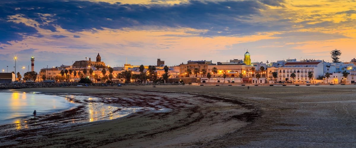 A coastal cityscape at dusk with illuminated buildings and a beach.