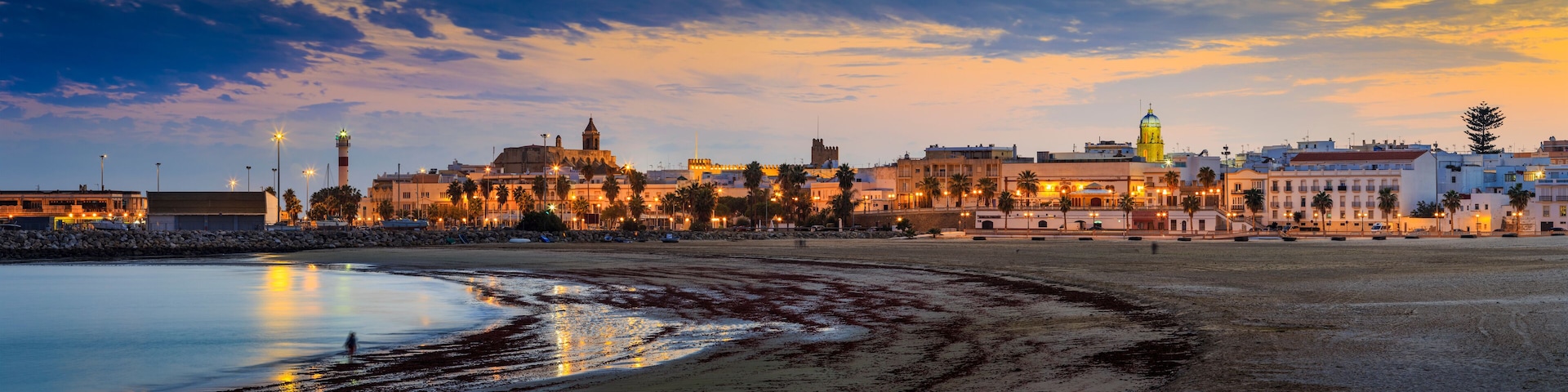 A coastal cityscape at dusk with illuminated buildings and a beach.