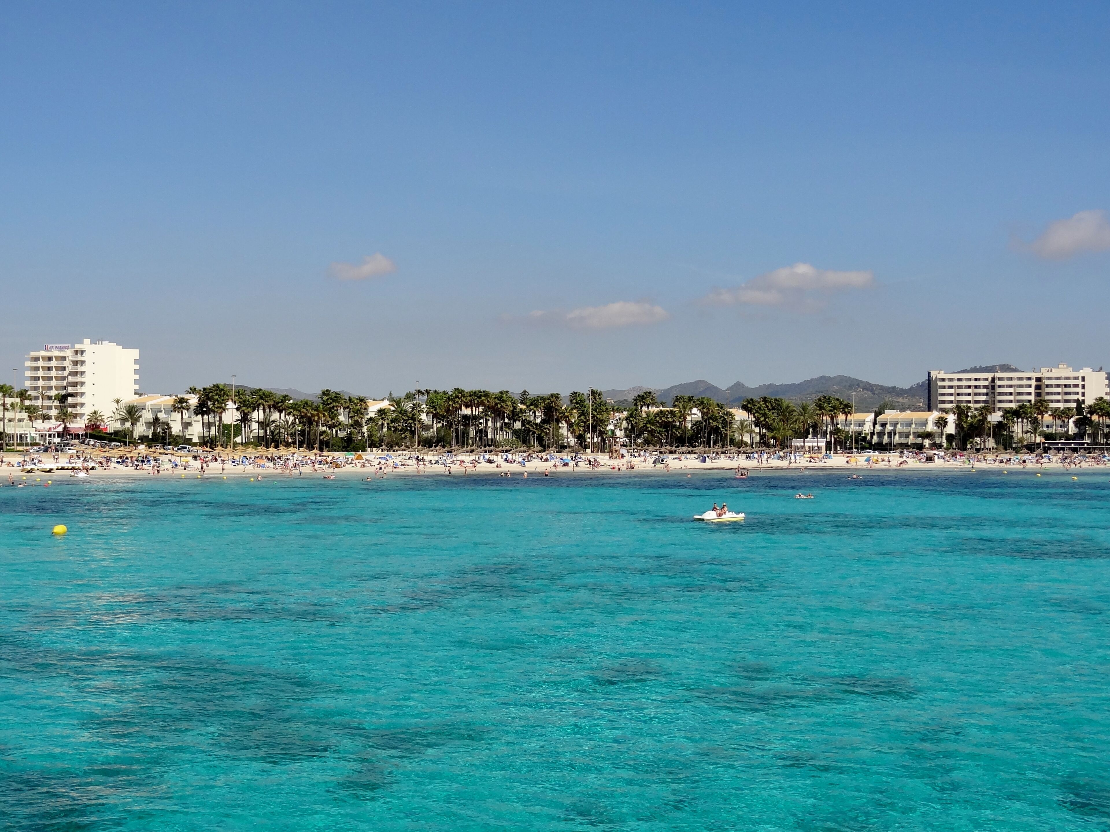 Una playa con agua turquesa cristalina, palmeras y edificios al fondo.