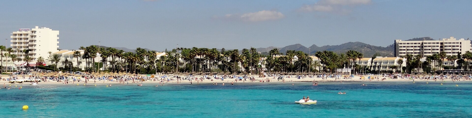 Una playa con agua turquesa cristalina, palmeras y edificios al fondo.