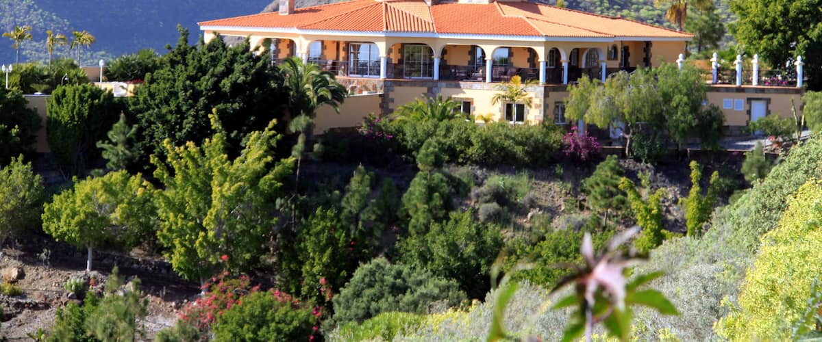 A large house with a red roof, surrounded by lush greenery and mountains in the background.