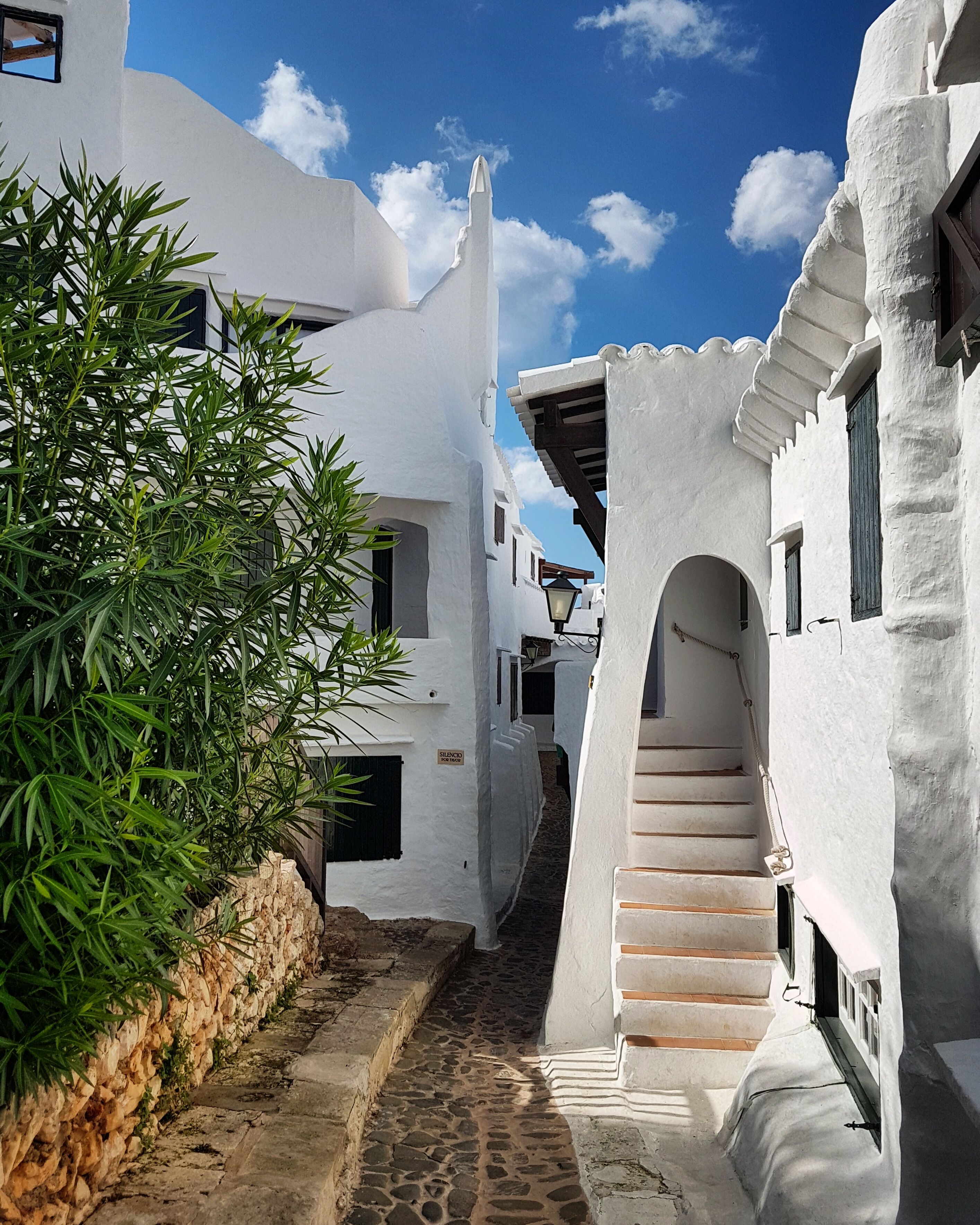 A narrow cobblestone street flanked by white buildings with black shutters, a stone wall, and a staircase leading to an upper level.