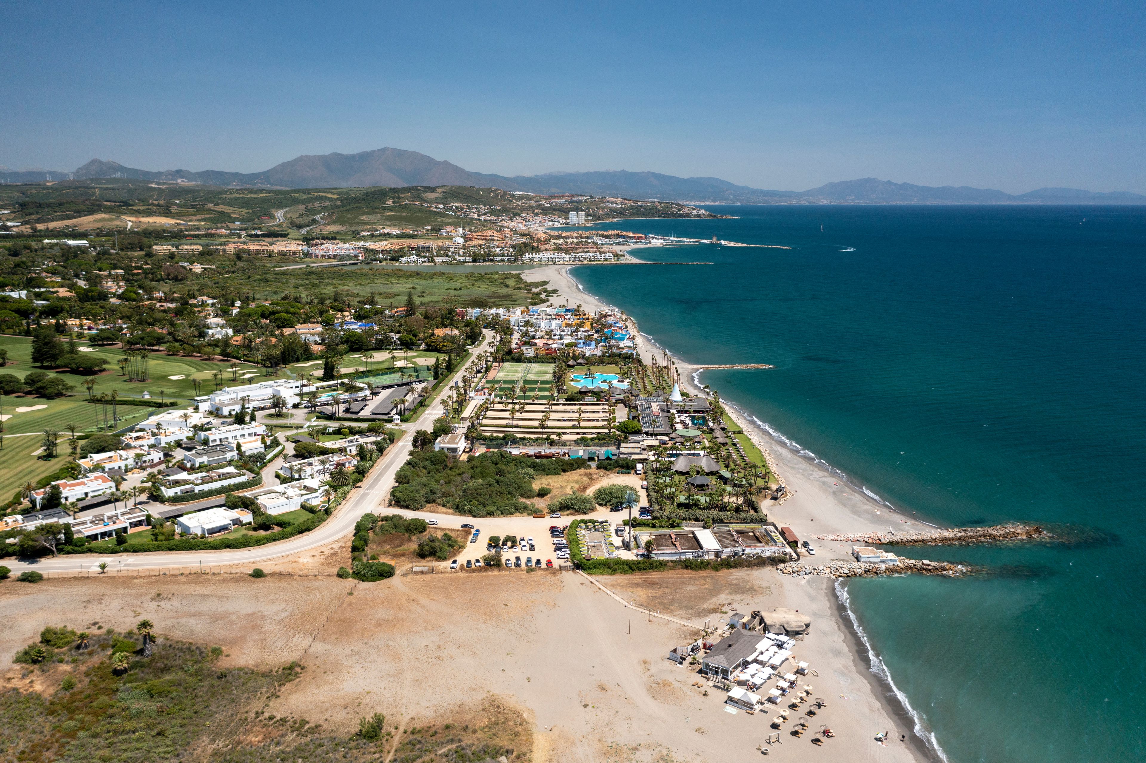 An aerial view of a coastal resort area with a beach, a golf course, and a mountainous backdrop.