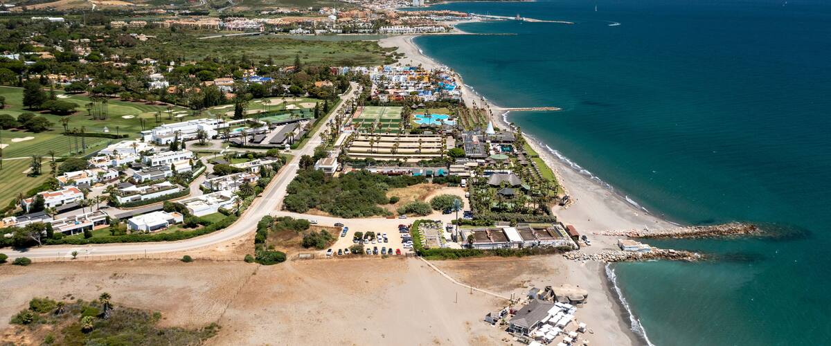 An aerial view of a coastal resort area with a beach, a golf course, and a mountainous backdrop.