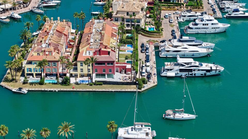 A marina with numerous yachts docked, surrounded by colorful buildings and palm trees.