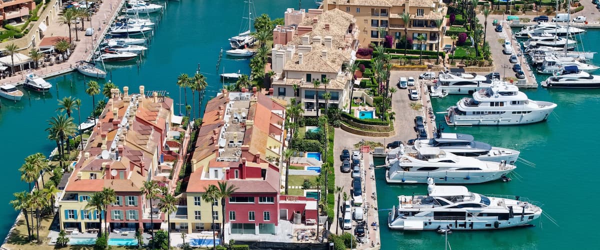 A marina with numerous yachts docked, surrounded by colorful buildings and palm trees.