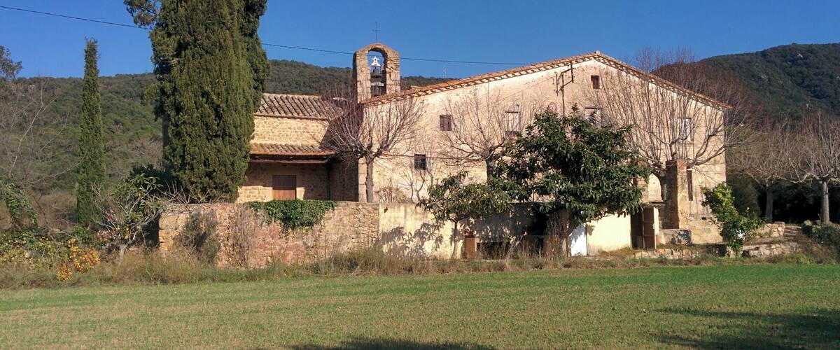A rustic stone building with a bell tower, surrounded by a grassy field and trees, set against a backdrop of hills.