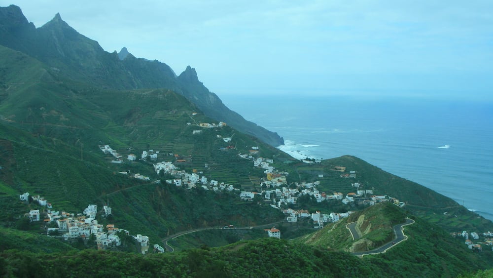 A coastal village nestled in green hills with a winding road and the ocean in the background.