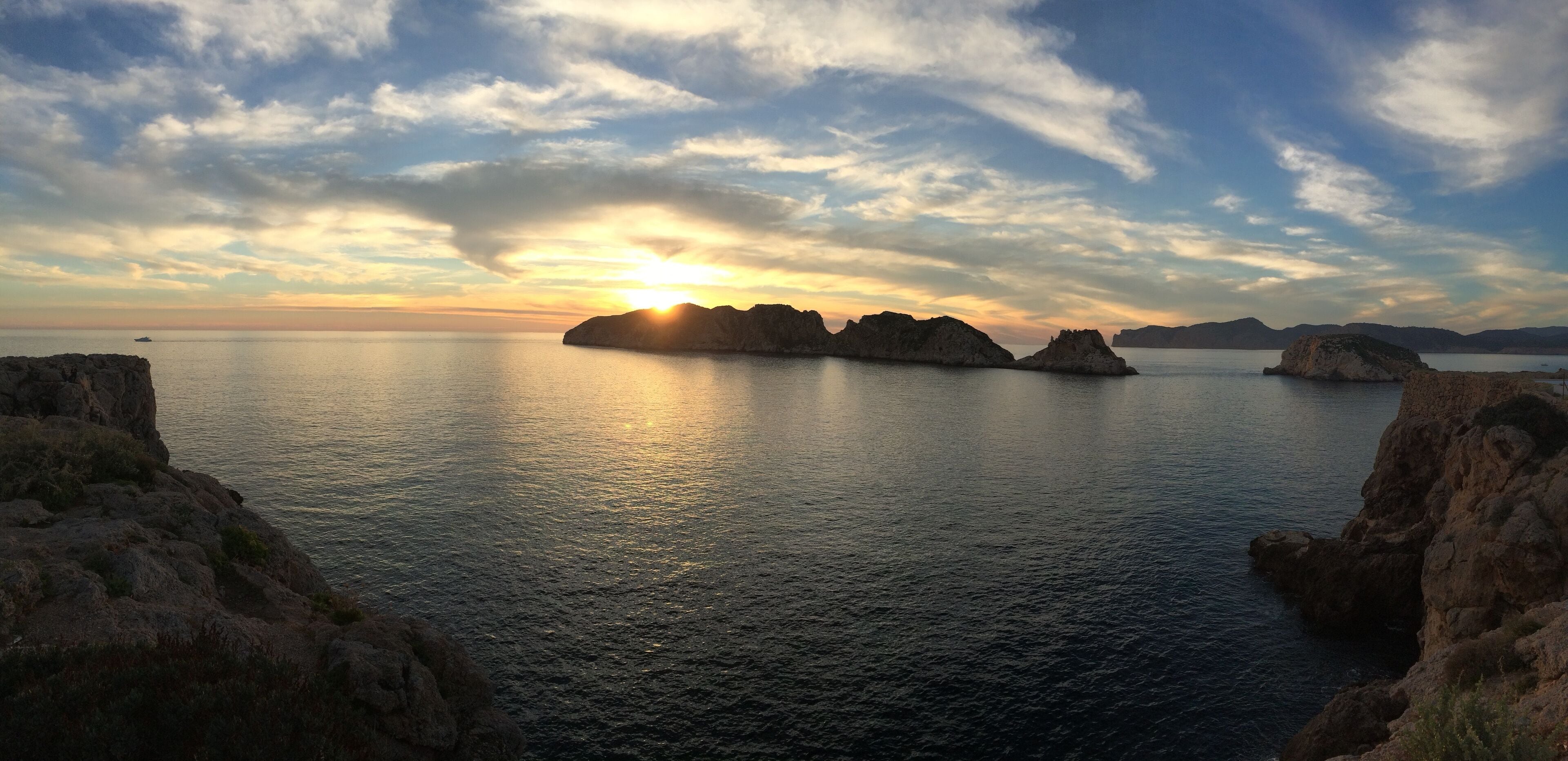 Un atardecer sobre un mar tranquilo con afloramientos rocosos y montañas distantes.