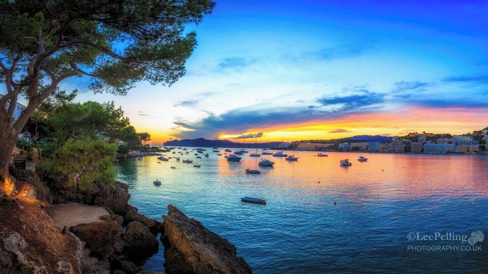 A coastal sunset with boats on calm waters, rocky shoreline, and buildings in the background.