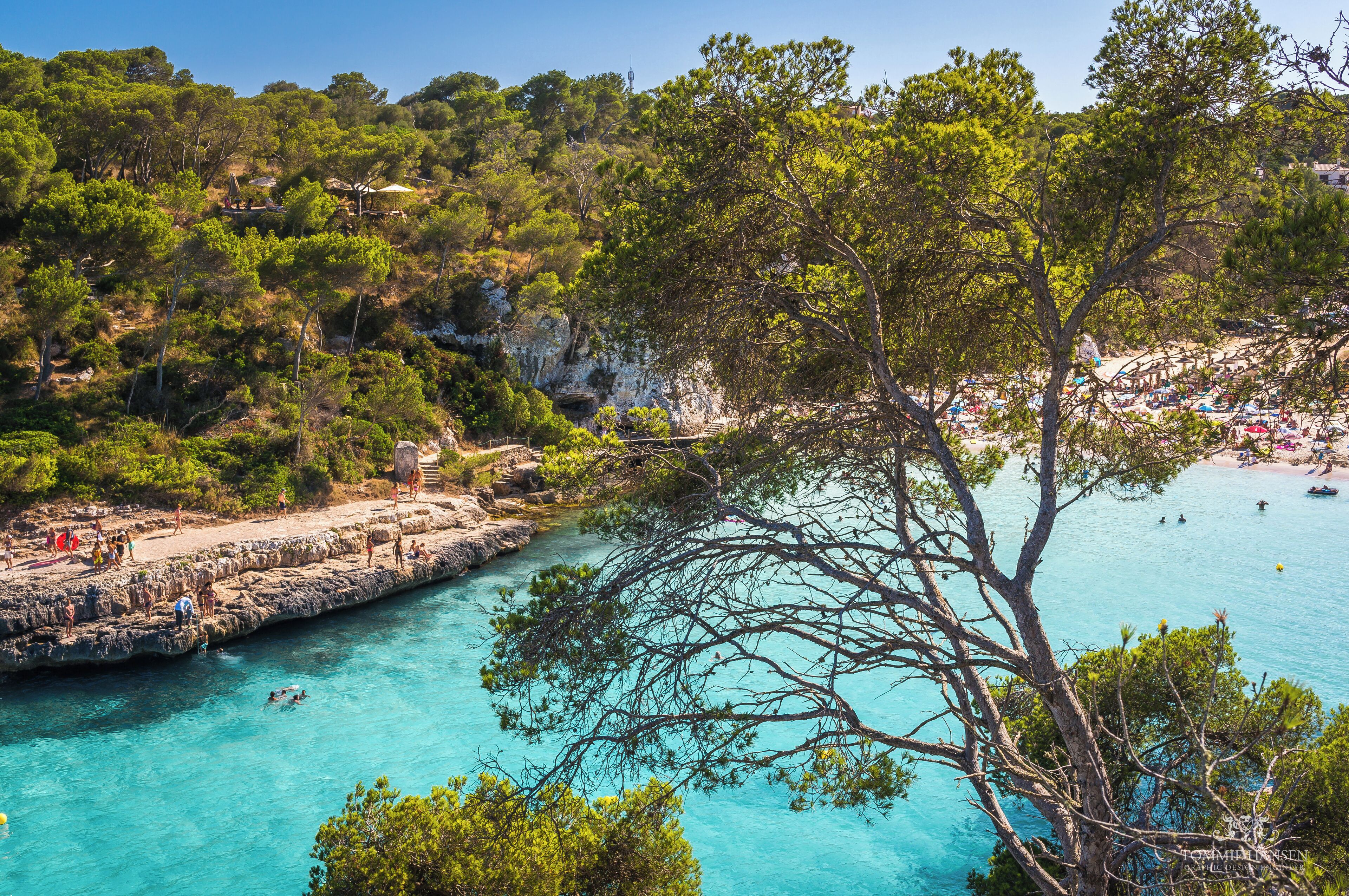 A coastal landscape with rocky cliffs, clear turquoise waters, and a beach area with people and boats.