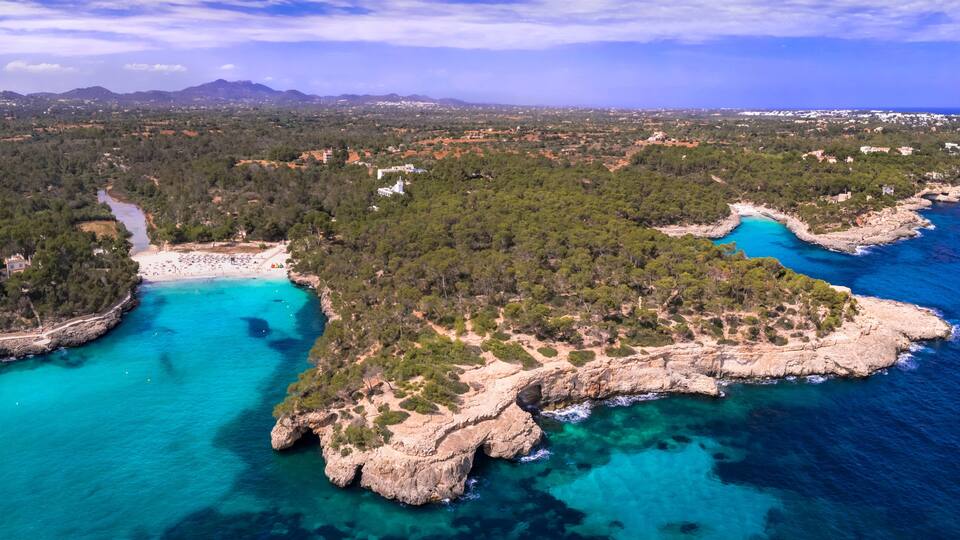 A coastal landscape with a beach, clear turquoise waters, rocky outcrops, and a dense forested area.