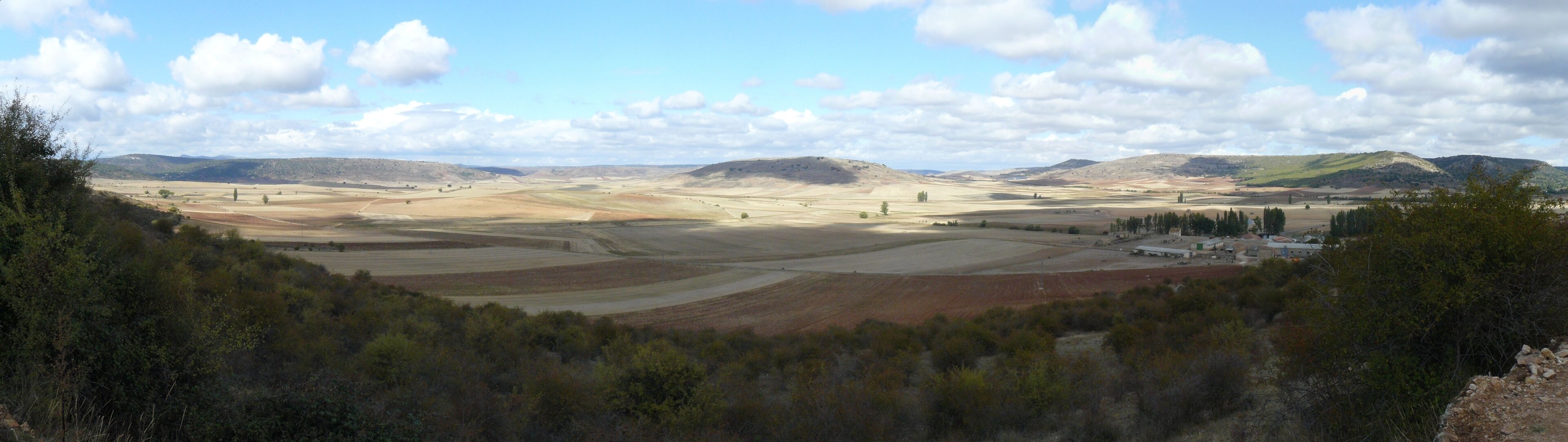 A vast landscape with rolling hills, fields, and a small cluster of buildings.