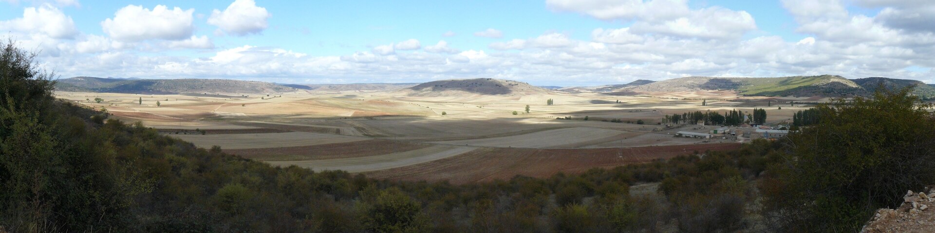 A vast landscape with rolling hills, fields, and a small cluster of buildings.