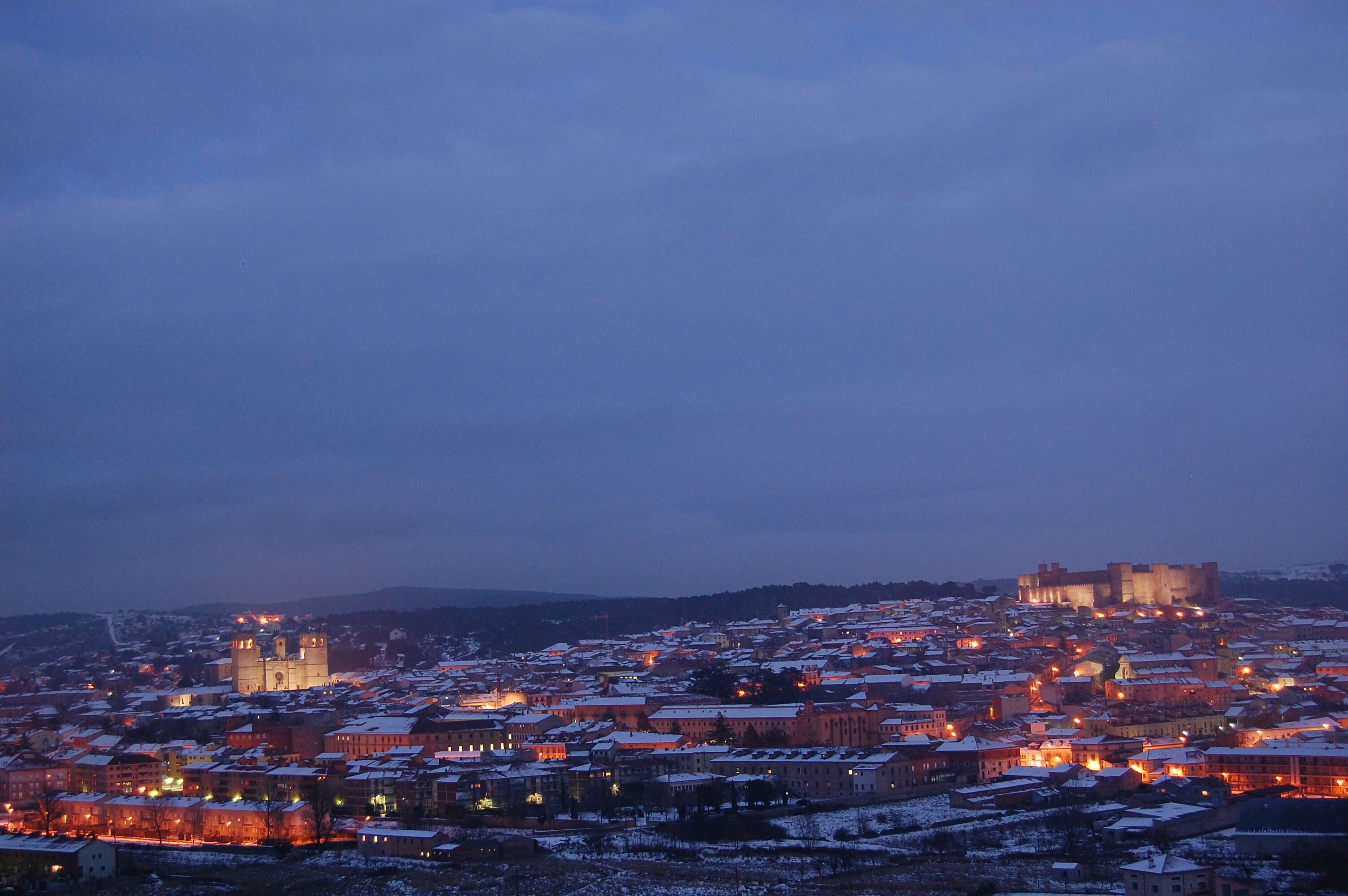 A cityscape at dusk with illuminated buildings and a prominent castle in the background.