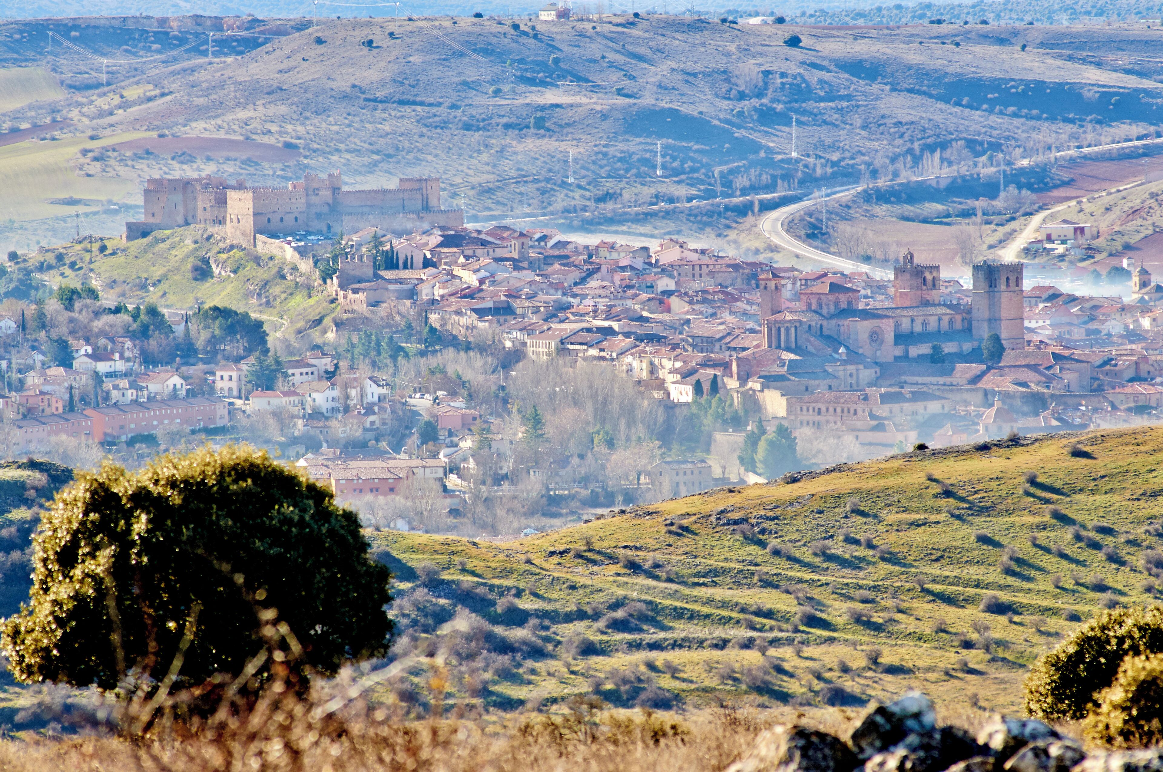 A historic town with a castle on a hill, surrounded by rolling hills and a river.