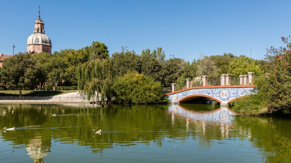 A bridge over a calm river, surrounded by lush greenery and a historic building with a dome in the background.