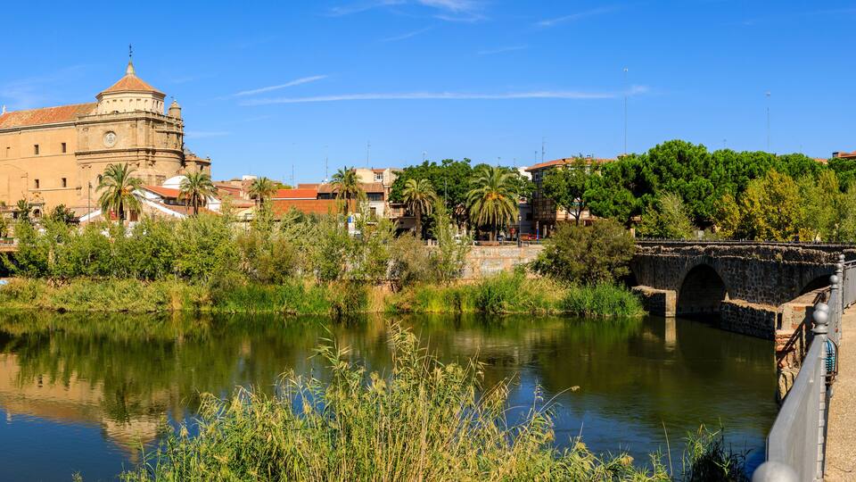 A river with a stone bridge, a historic building with a dome, and lush greenery.