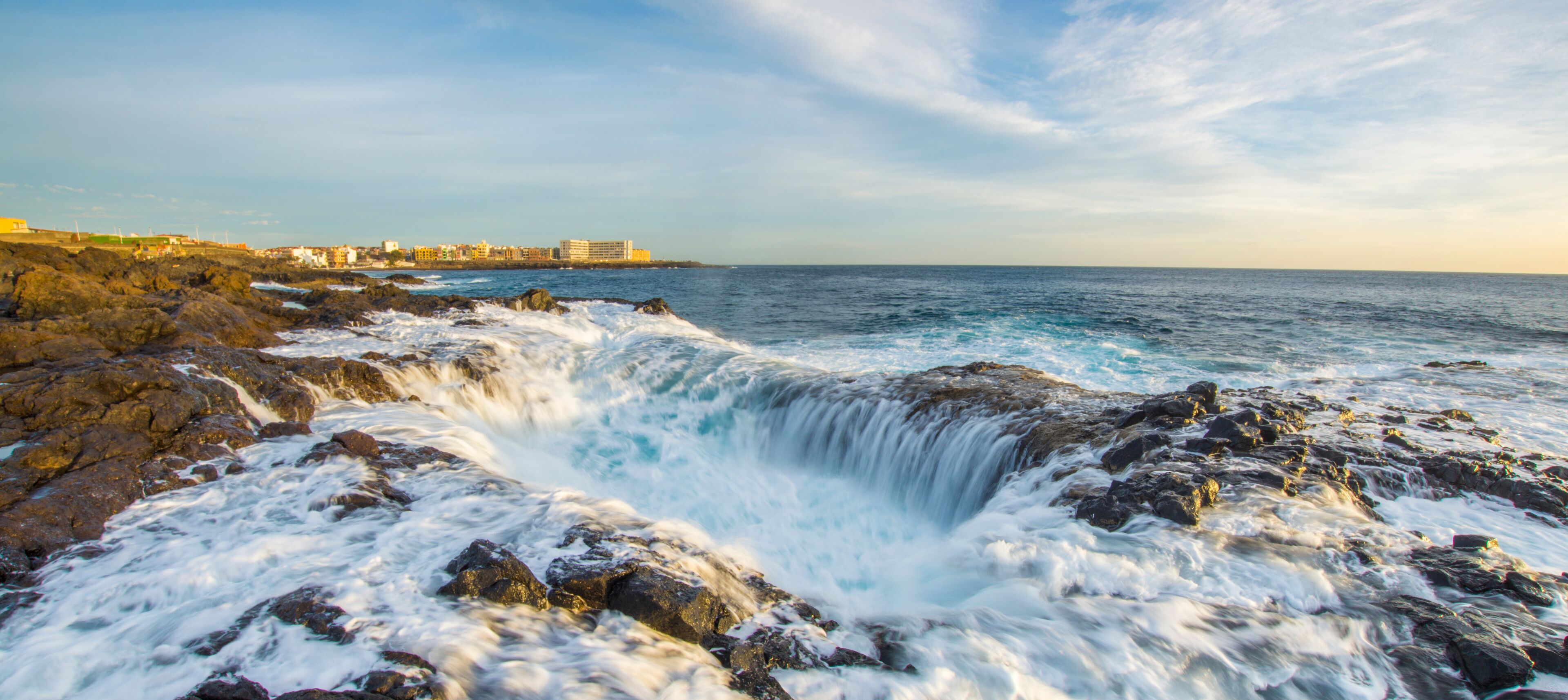A rocky coastline with waves crashing against the rocks and a distant view of buildings along the shore.