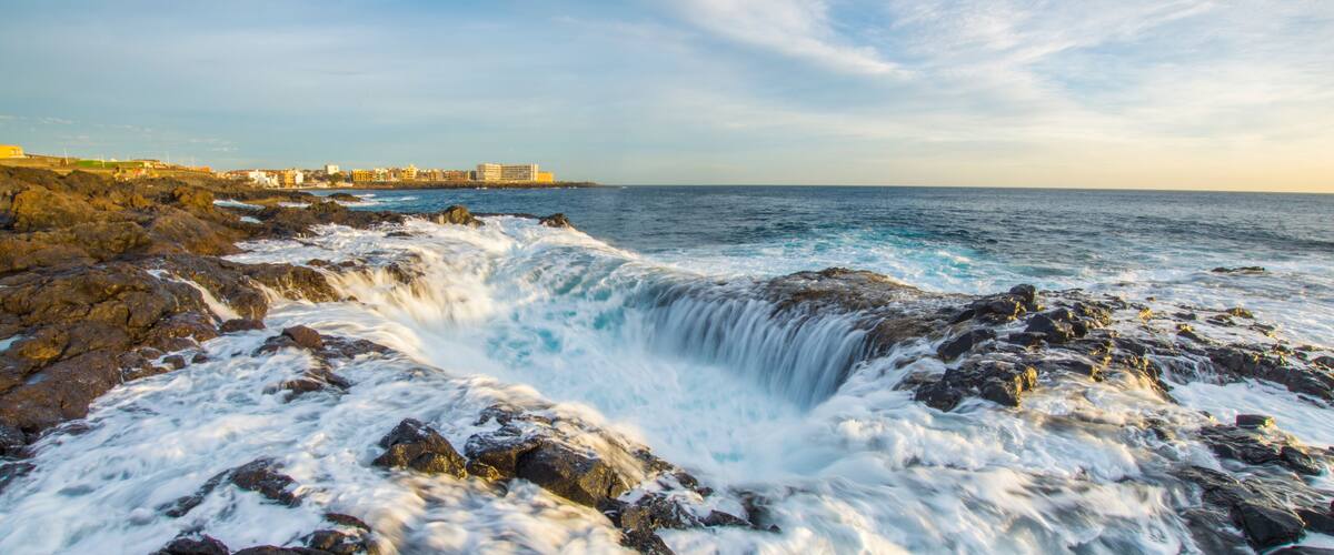 A rocky coastline with waves crashing against the rocks and a distant view of buildings along the shore.