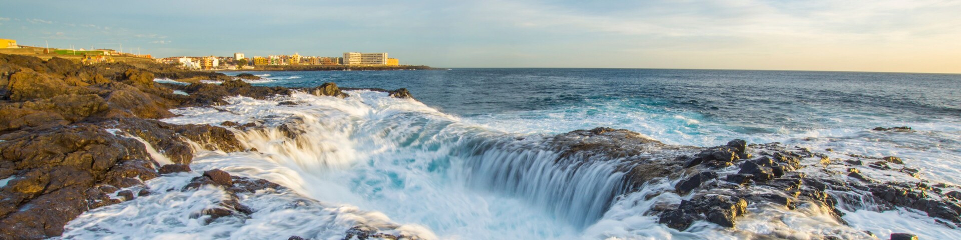 A rocky coastline with waves crashing against the rocks and a distant view of buildings along the shore.