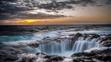 Paisagem costeira com cachoeira, ondas do oceano e céu ao pôr do sol.