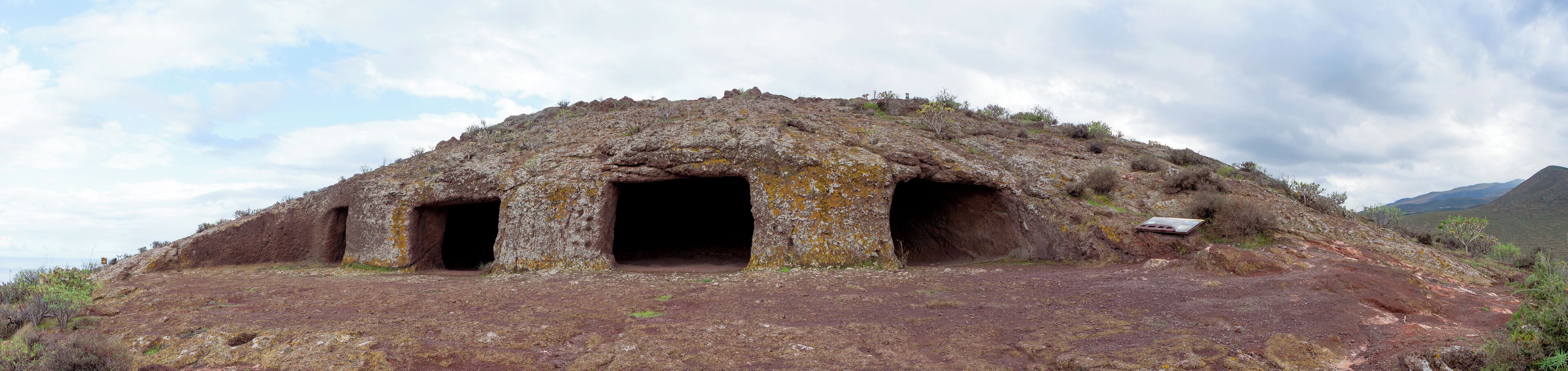 A large, ancient, rock-covered structure with three openings, surrounded by a barren landscape and mountains in the background.