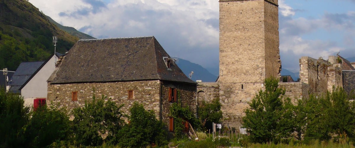 A historic stone church with a tall bell tower, surrounded by lush greenery and mountains in the background.