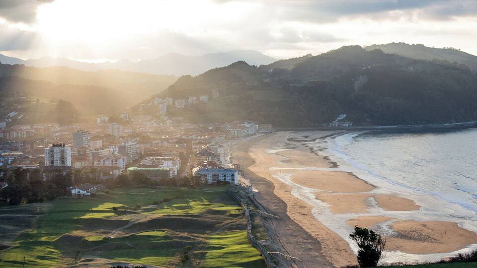 A coastal city with a beach, buildings, and rolling hills in the background.
