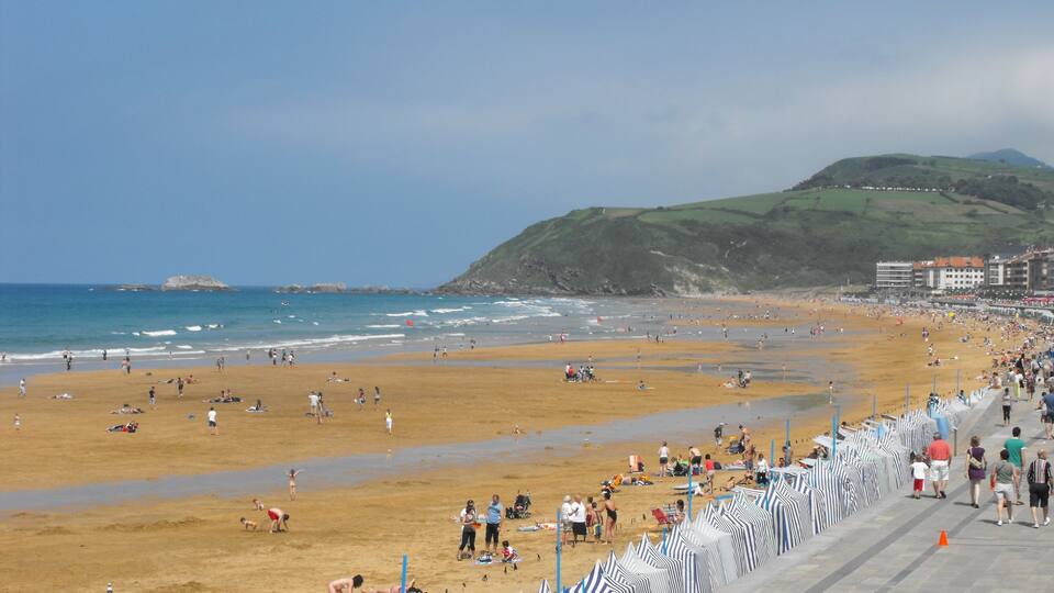A crowded beach with people sunbathing and swimming, a rocky outcrop in the water, and a hill in the background.