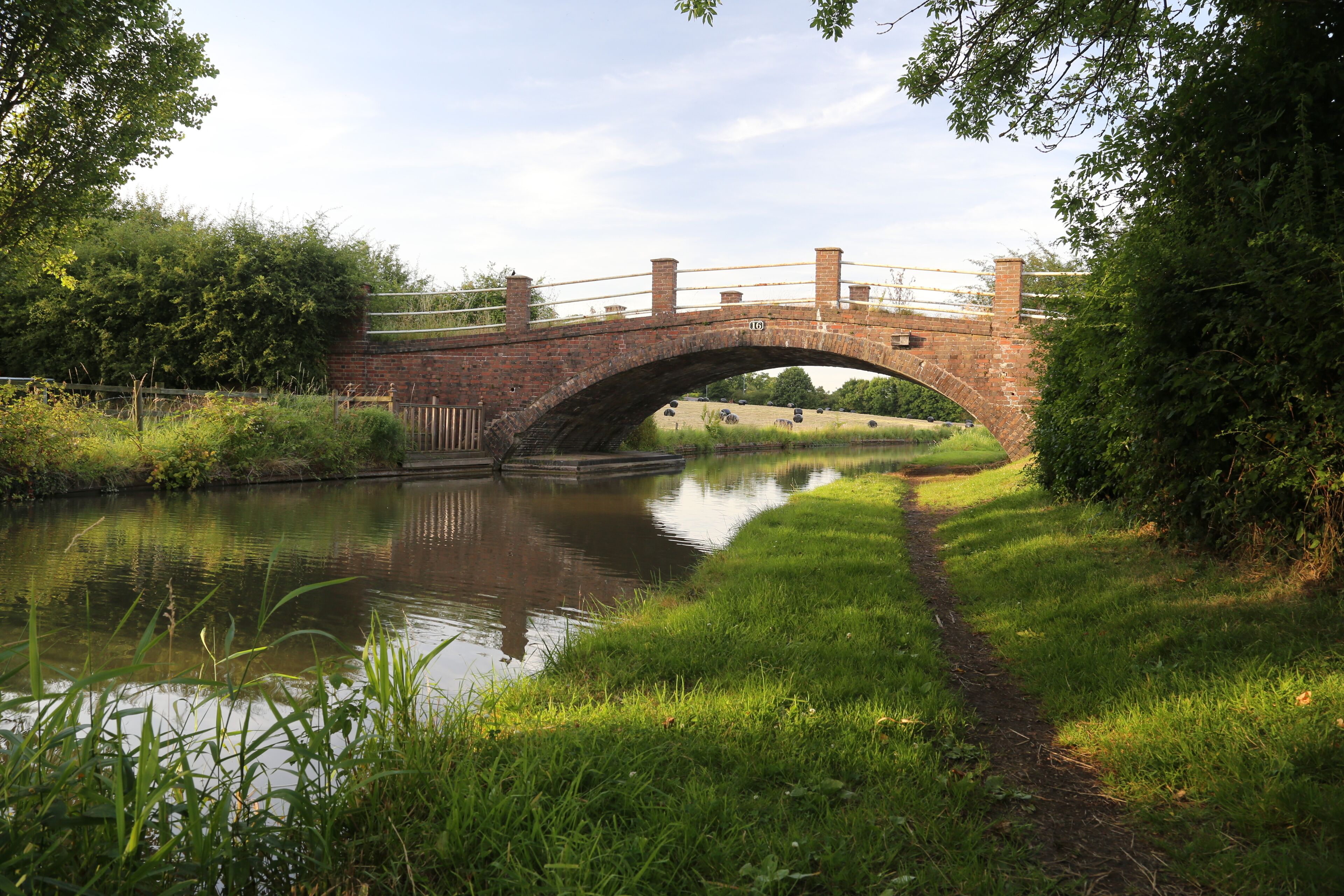 A brick bridge over a calm river, surrounded by lush greenery and a grassy path.