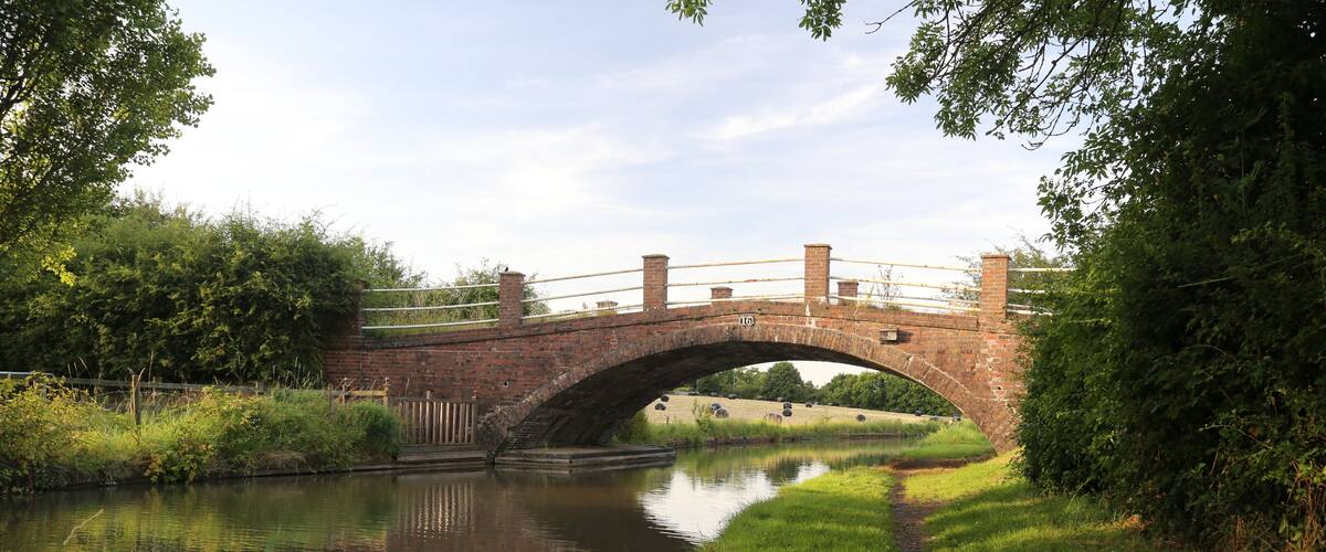 A brick bridge over a calm river, surrounded by lush greenery and a grassy path.