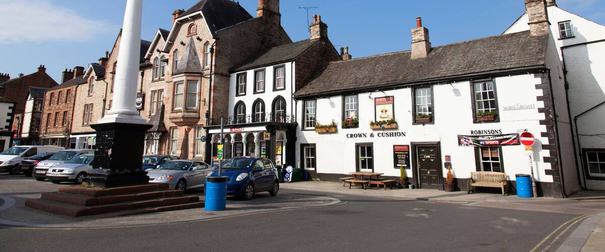 A town square with a prominent white monument, a pub named 'Crown & Cushion', and several parked cars.