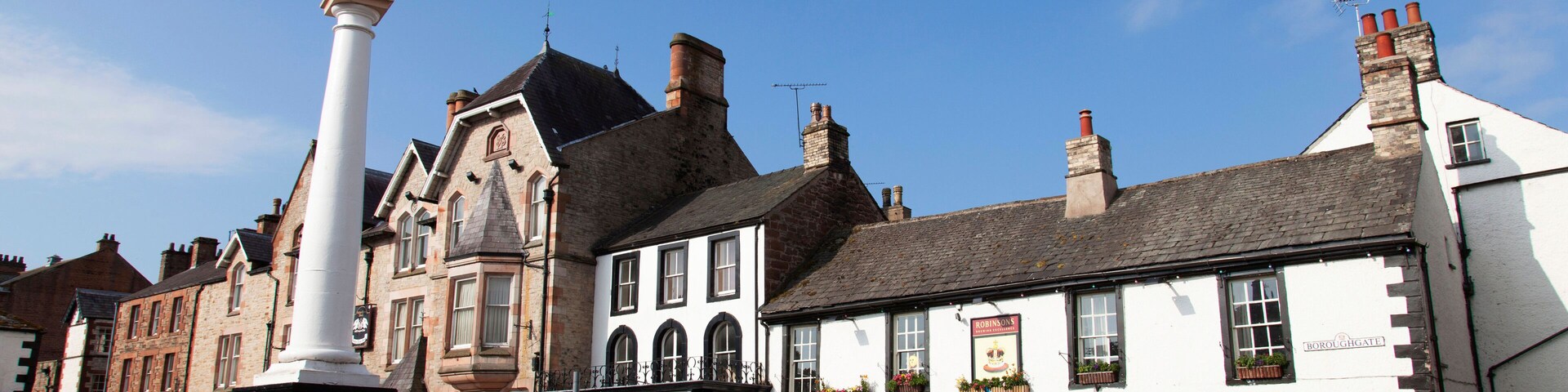 A town square with a prominent white monument, a pub named 'Crown & Cushion', and several parked cars.