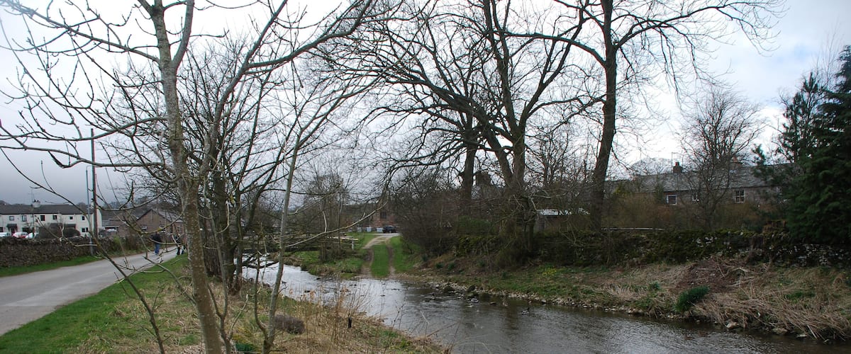 A narrow river with a path alongside it, surrounded by trees and a rural landscape.