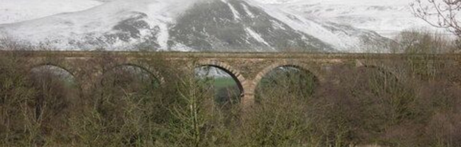 Un puente de piedra sobre un río, rodeado de campos verdes y colinas nevadas.