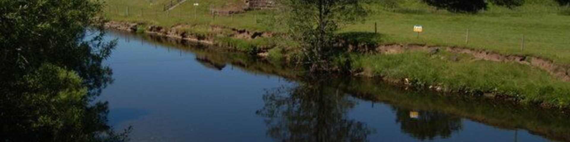 A calm river with a small house and grassy fields in the background.