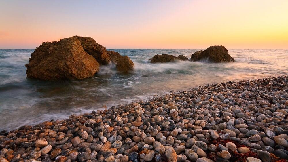 Une plage de galets avec de gros rochers dans l’eau et un ciel au coucher du soleil.