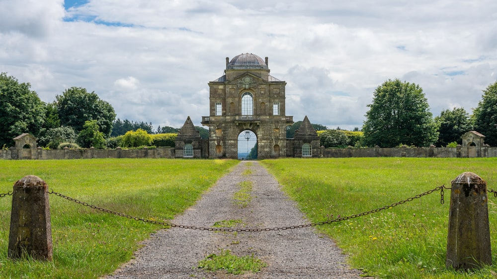 Un bâtiment historique en pierre, doté d’une grande entrée en arc de cercle, entouré d’un champ verdoyant et accessible par un chemin de gravier.