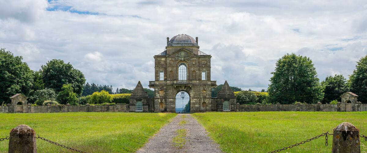 A historic stone building with a large arched entrance, surrounded by a grassy field and a gravel path leading to it.