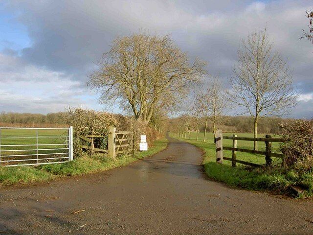 A rural road with a metal gate and wooden fence on the left, leading to a grassy field.