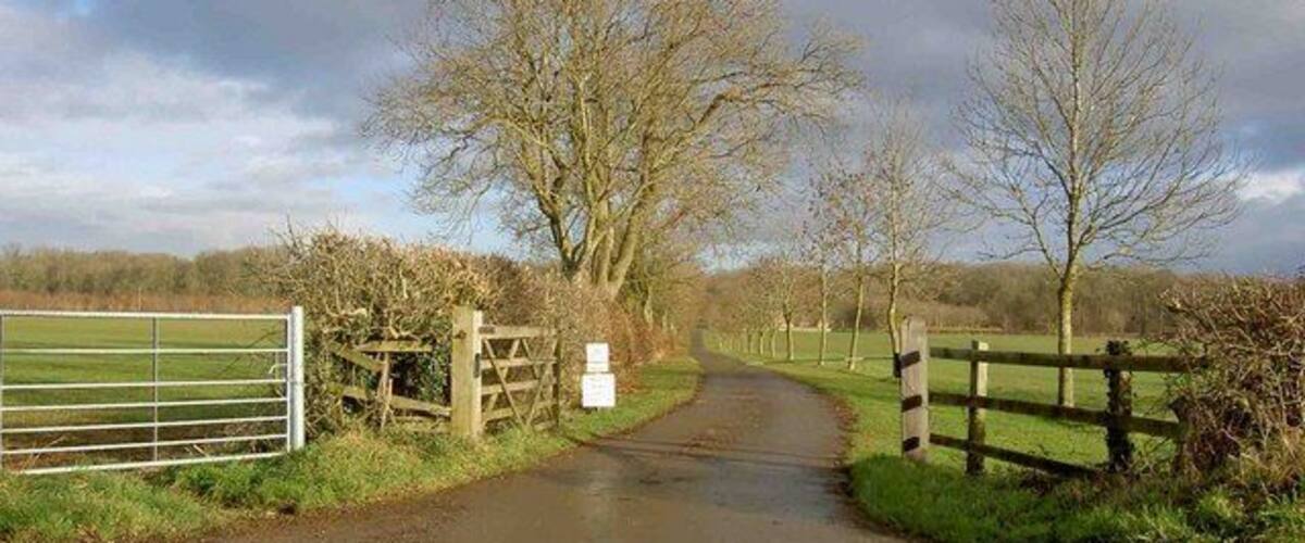 A rural road with a metal gate and wooden fence on the left, leading to a grassy field.